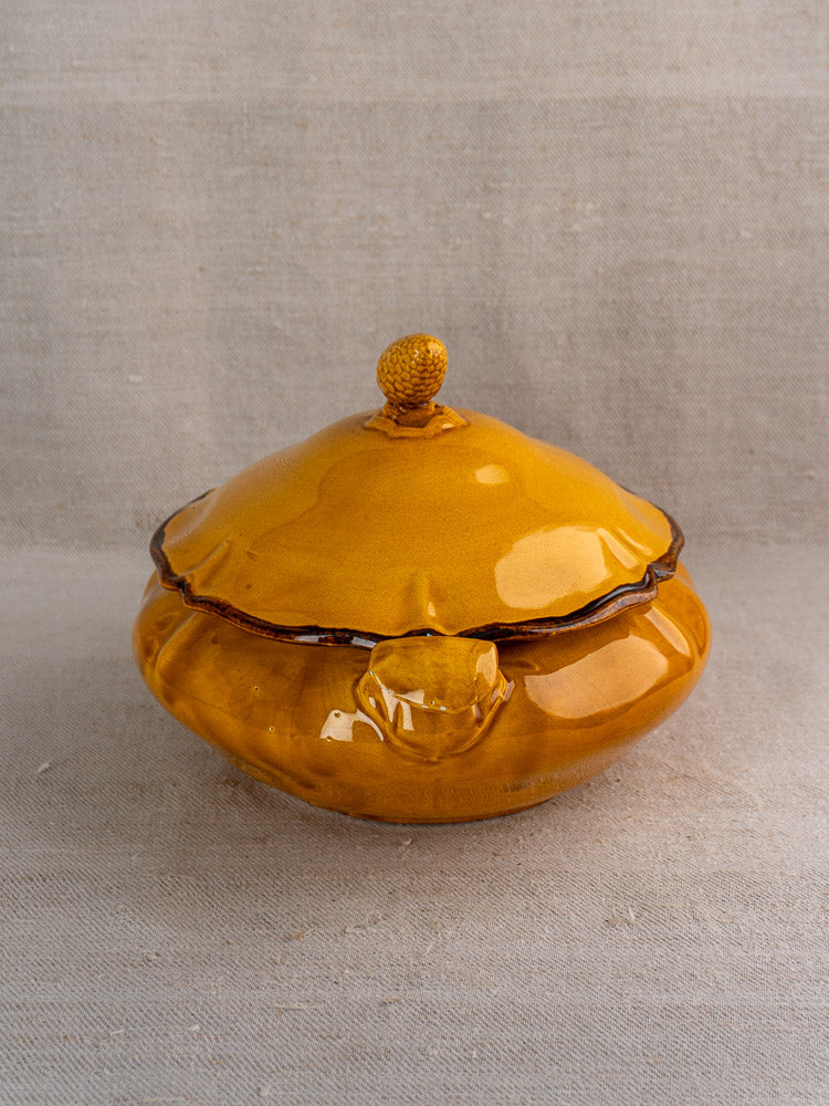 Vintage French Haute Provence faience tureen with yellow glaze, scalloped edges, pine cone finial, on neutral fabric.