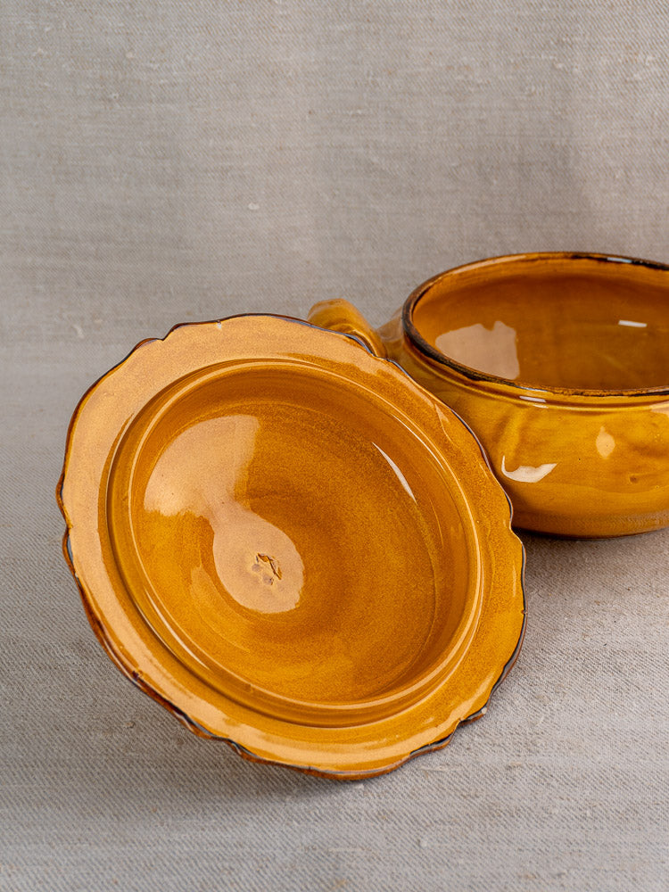 Pair of yellow-brown ceramic tureens with scalloped edges and pine cone finials on neutral fabric.