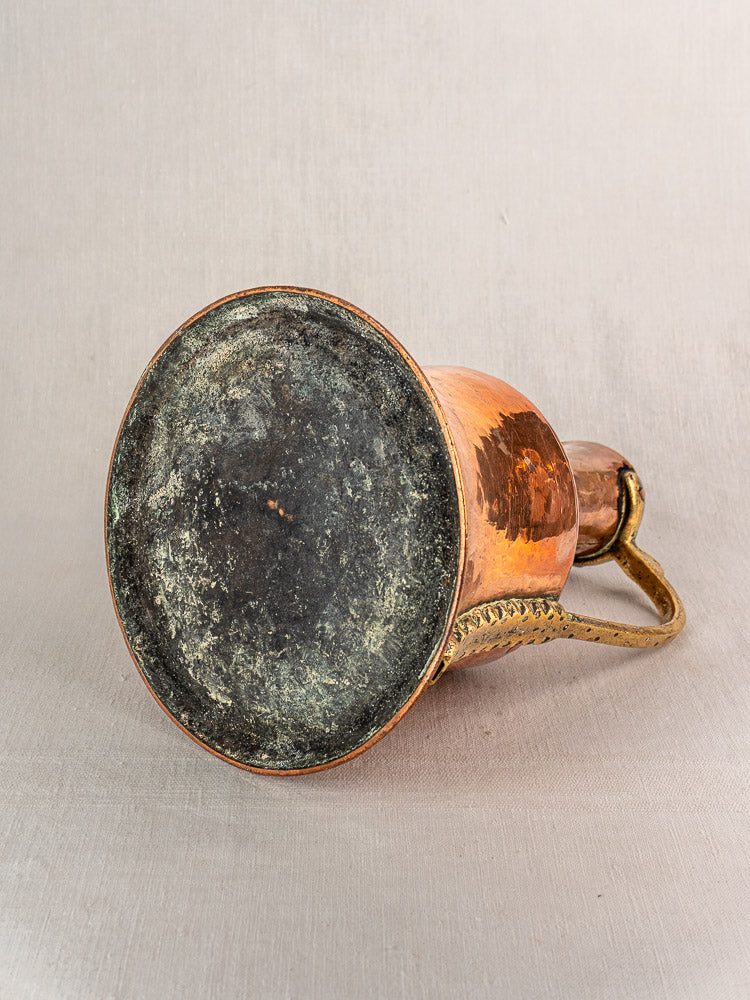Antique French copper water jug with brass handle showing weathered base on beige fabric background.