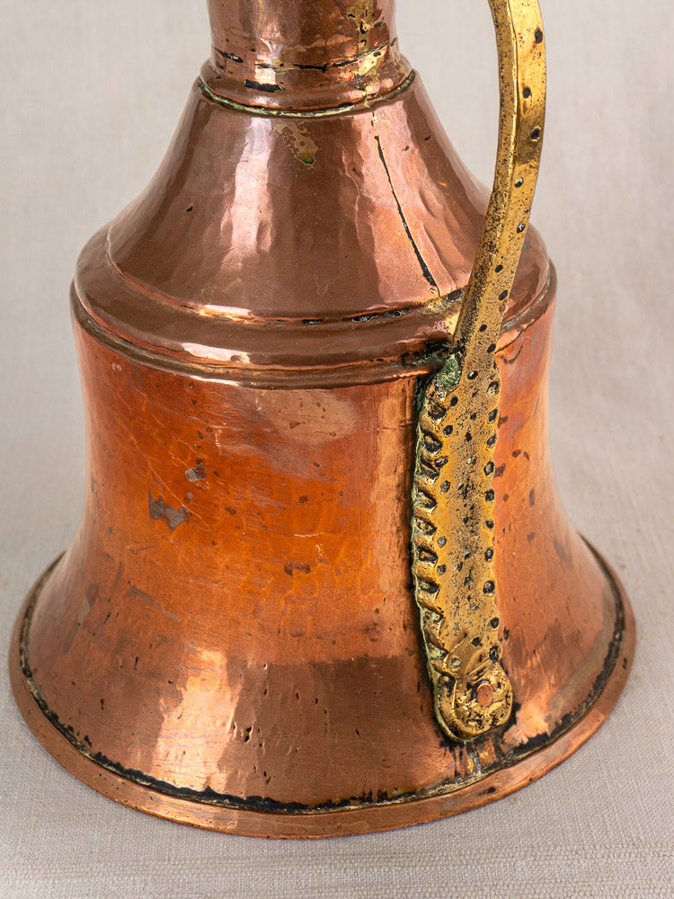 Antique French copper water jug with wide base, golden brass handle, and aged patina on neutral fabric background.