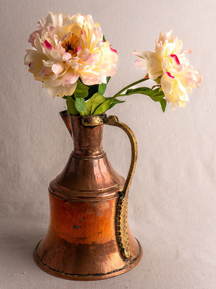 Antique French copper water jug with brass handle, filled with pale yellow peonies, on a beige background.