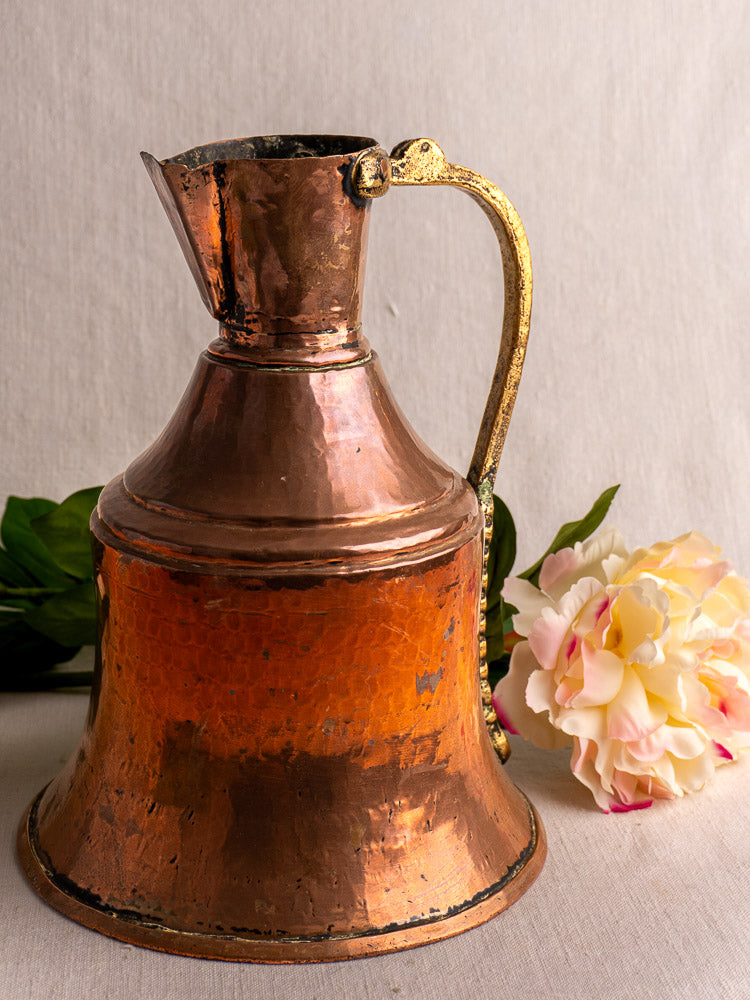 Antique French copper water jug with brass handle, set before green leaves and a pink artificial flower.