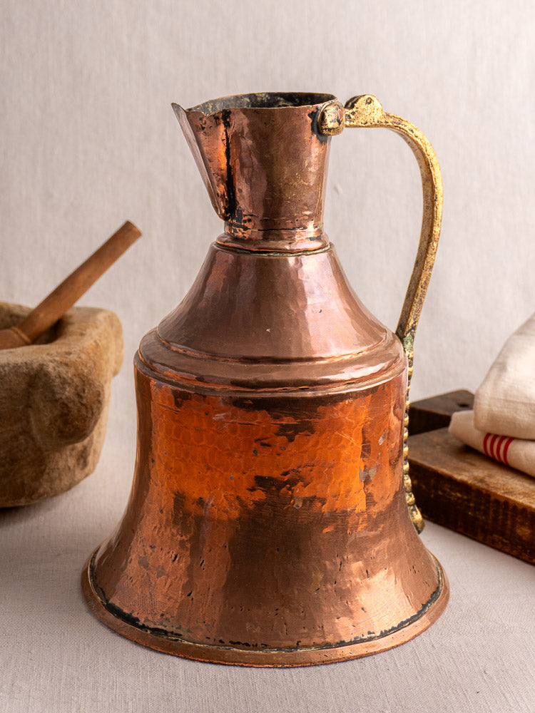Antique French copper water jug with brass handle on a light surface, stone mortar and red-striped linens behind.