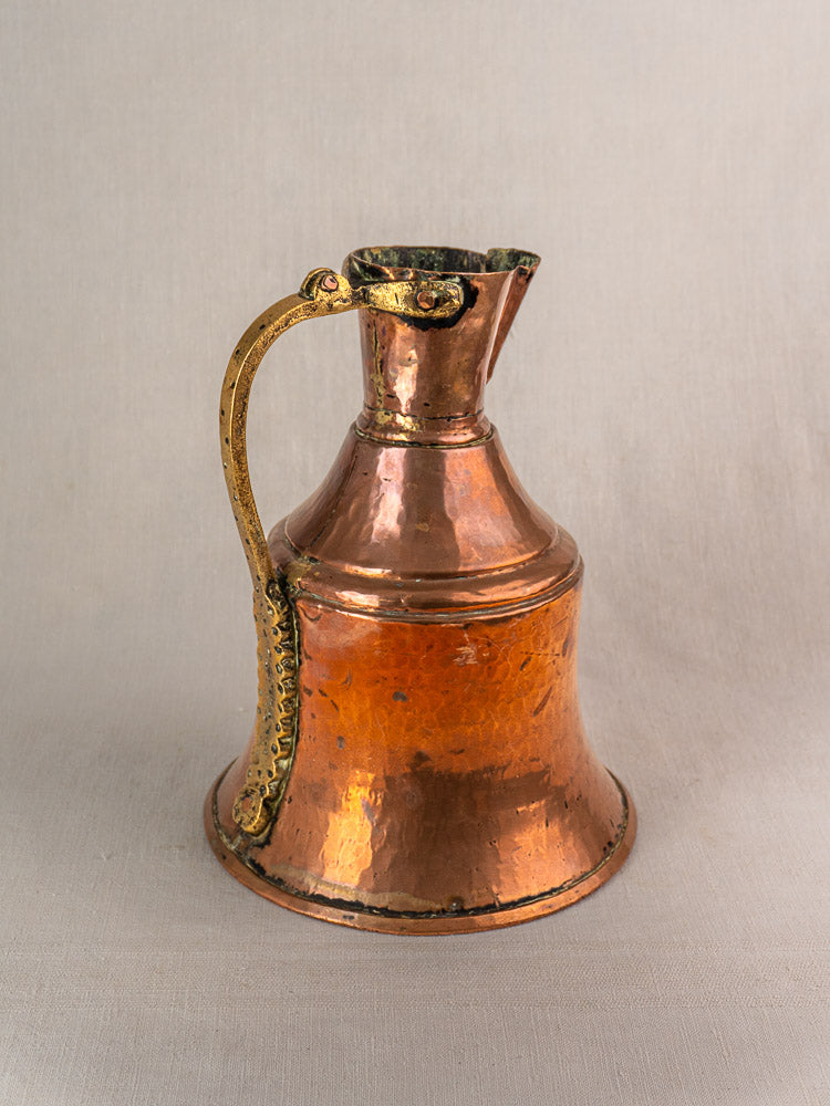 Antique French copper water jug with wide base, narrow neck, brass handle; set against plain light fabric backdrop.