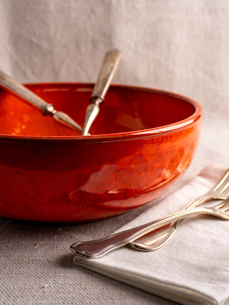 Antique French earthenware serving bowl with vibrant orange glaze, silver utensils, and forks on neutral fabric.