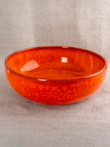 Antique French earthenware serving bowl with vibrant orange glaze on light beige textured fabric background.