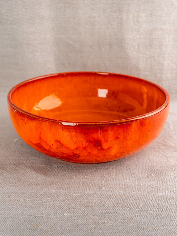 Antique French earthenware serving bowl with vibrant orange glaze on textured beige fabric background. Mid 20th century.