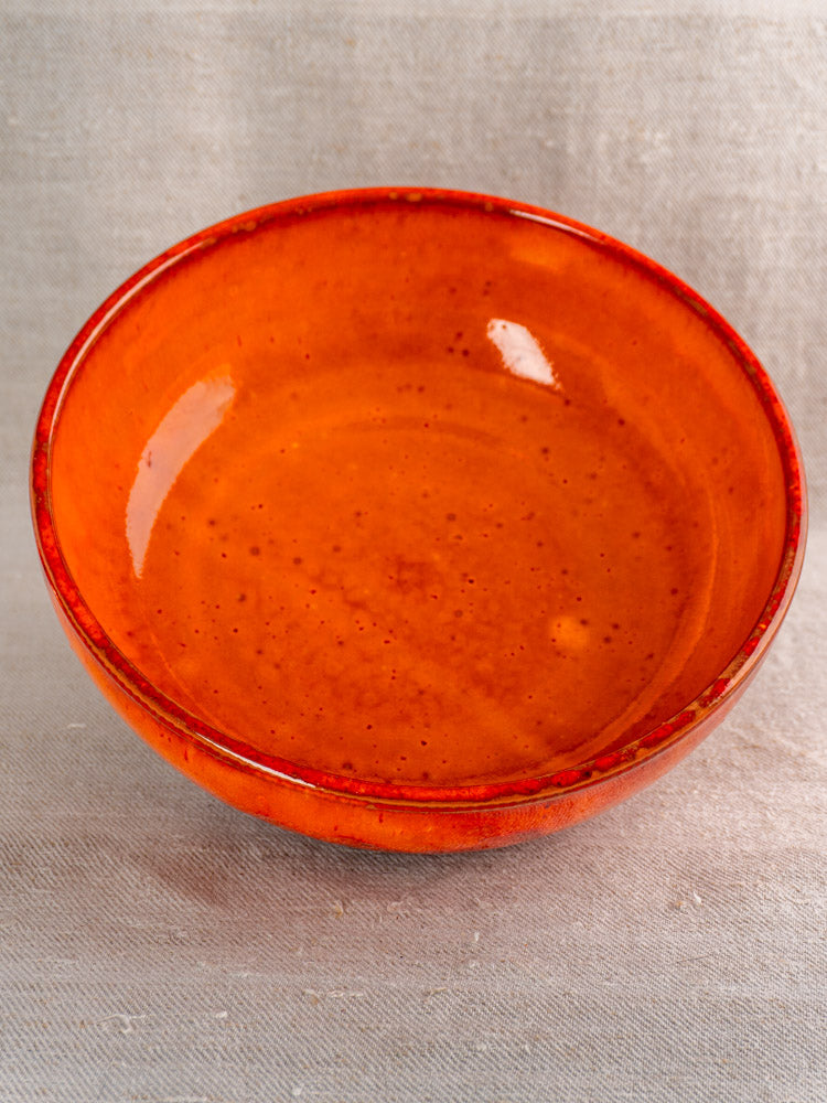Antique French earthenware serving bowl with vibrant orange glaze and glossy finish on textured beige fabric background.