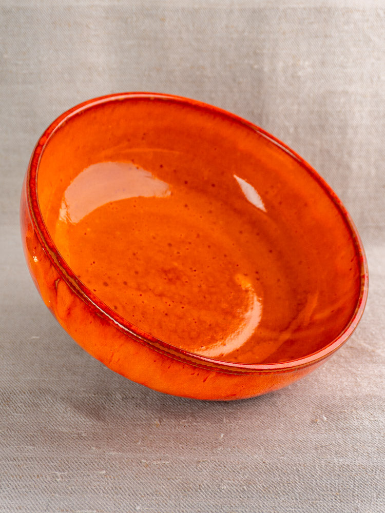 Mid-century French earthenware serving bowl with vibrant orange glaze, subtle speckles, on beige fabric surface.