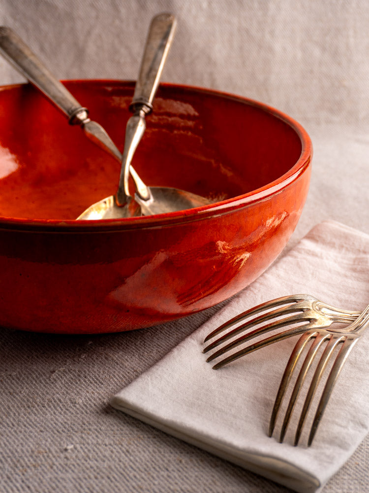 Antique French earthenware serving bowl with vibrant orange glaze on light beige textured fabric background.