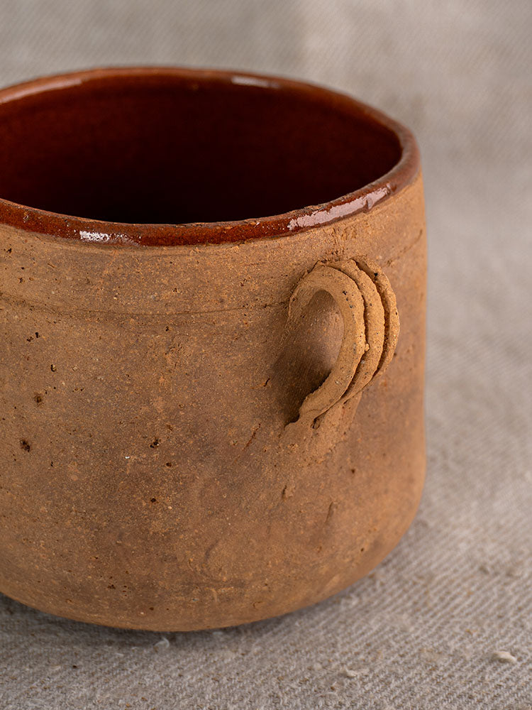 Late 19th-century French confit pot with rustic clay body, looped handle, brown glazed interior on gray fabric background.