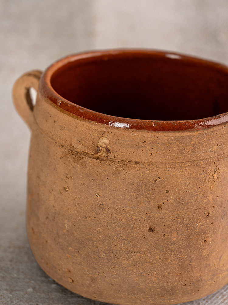 Antique French confit pot, late 19th century, rustic brown exterior, small handle, glazed interior, on a light surface.