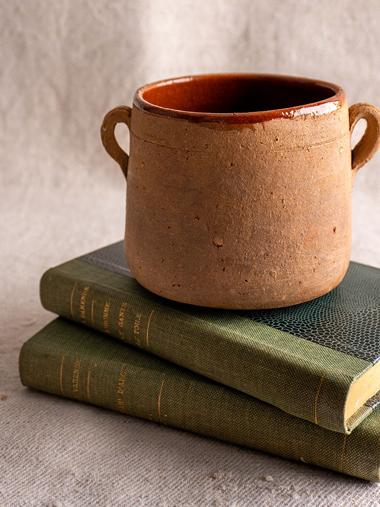 Antique French confit pot with brown glazed interior on stacked green hardcovers against a neutral textured backdrop.