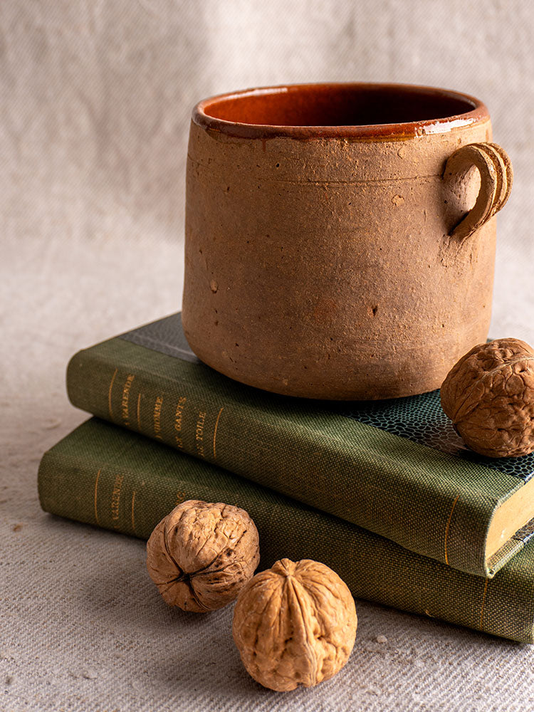 Antique French confit pot with brown glazed interior atop green books, walnuts nearby on a textured beige surface.