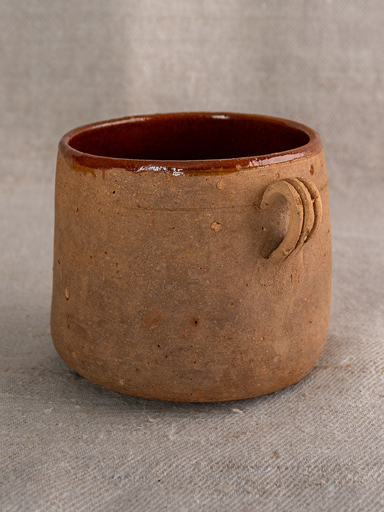 Antique French confit pot with textured brown exterior, glazed interior, and decorative swirl on neutral fabric background.
