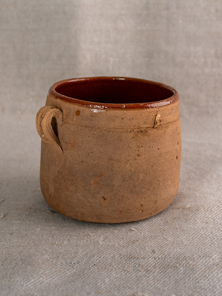 Antique French confit pot with brown glazed interior and single handle on textured gray fabric.