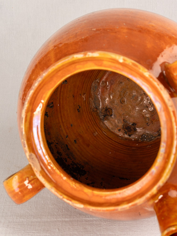 Antique French glazed earthenware cruche with handles, honey-brown finish, dark interior residue, on light fabric surface.
