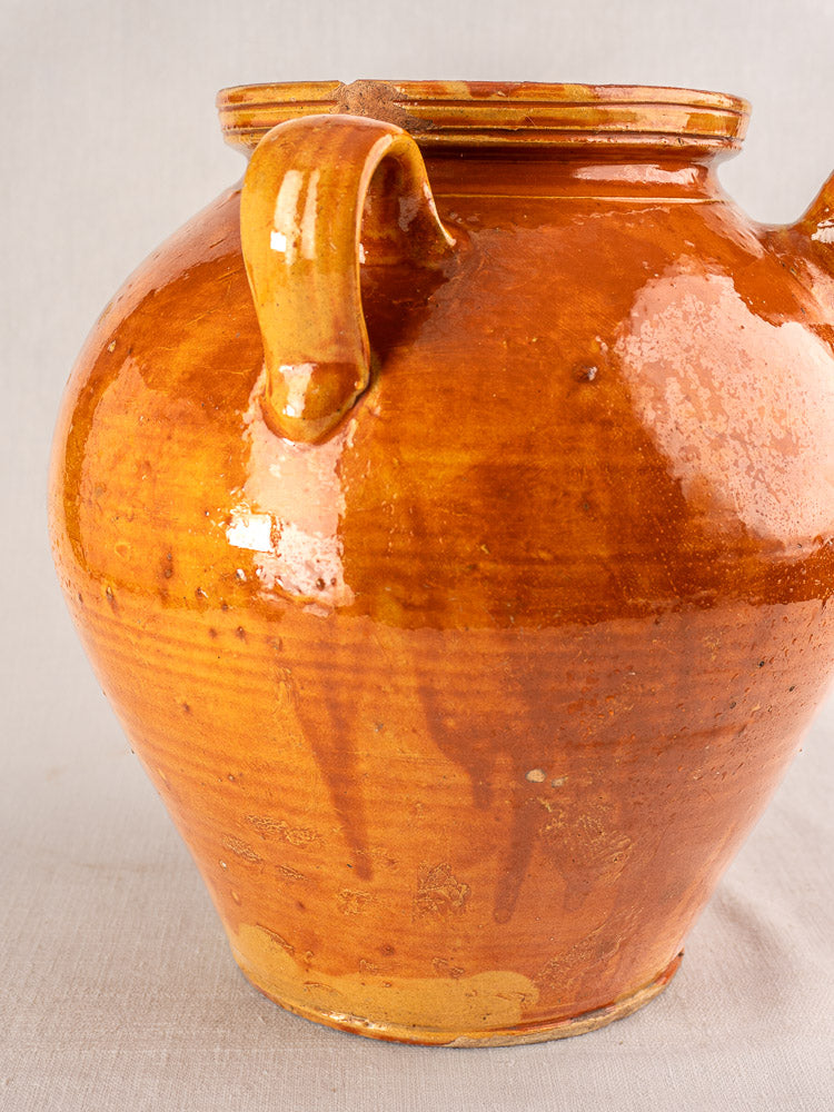 Antique French glazed earthenware cruche with honey-brown finish, two handles, glaze drips; neutral gray background.