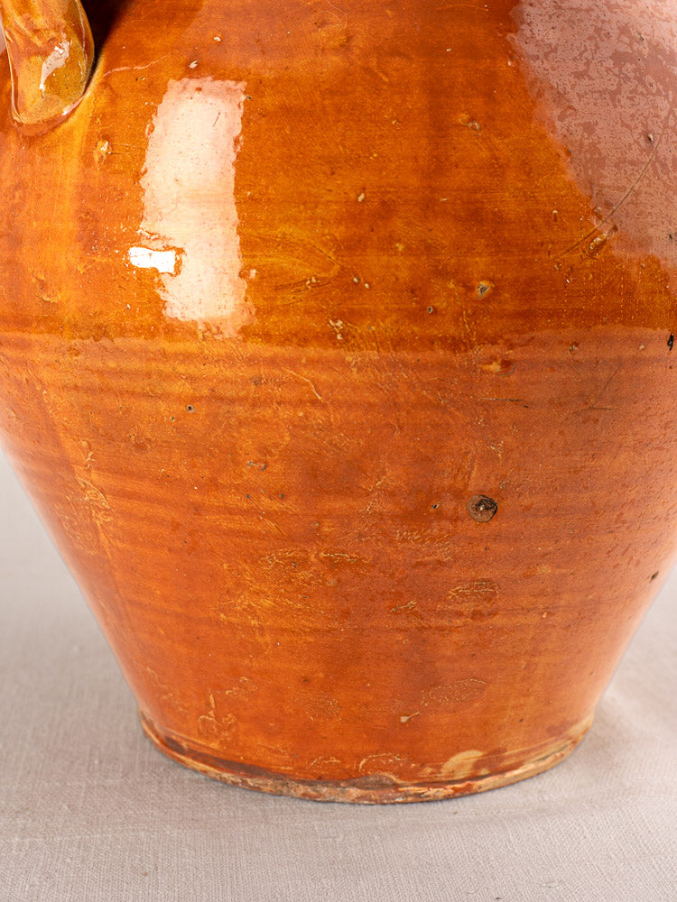 Antique French cruche with orange-brown glazed earthenware, handle, and textured surface on light fabric.