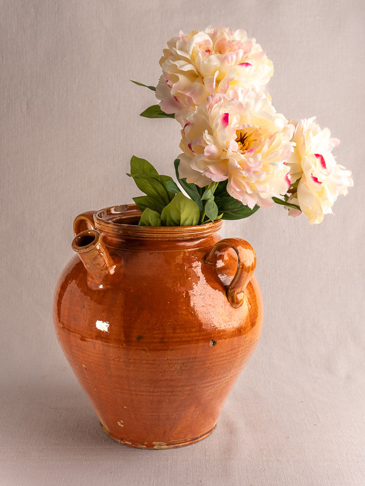 Antique French glazed earthenware cruche with dual handles, honey-brown finish, and faux peonies on neutral fabric.