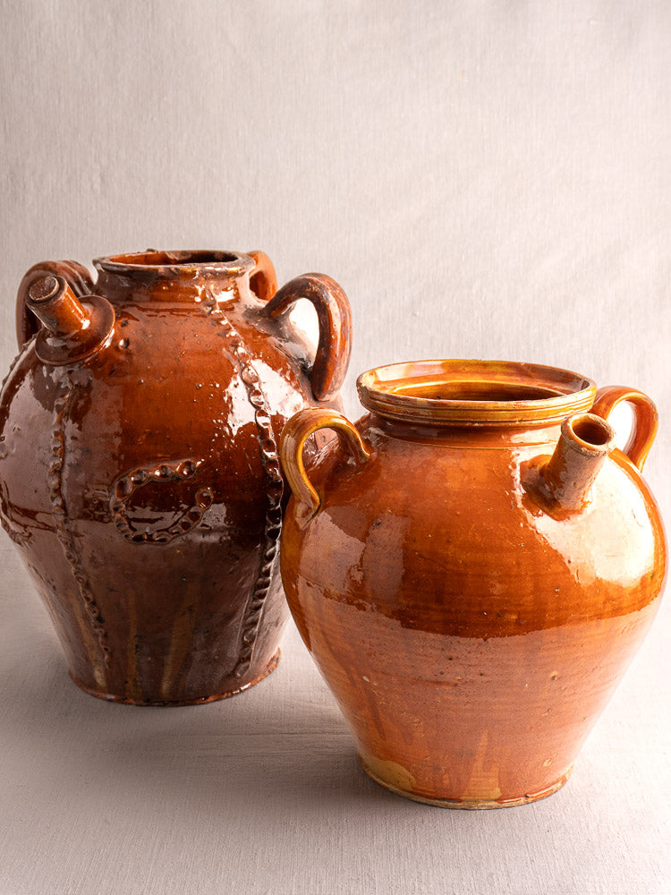 Pair of antique French glazed earthenware cruche jugs with three handles, honey-brown finish, on light surface.