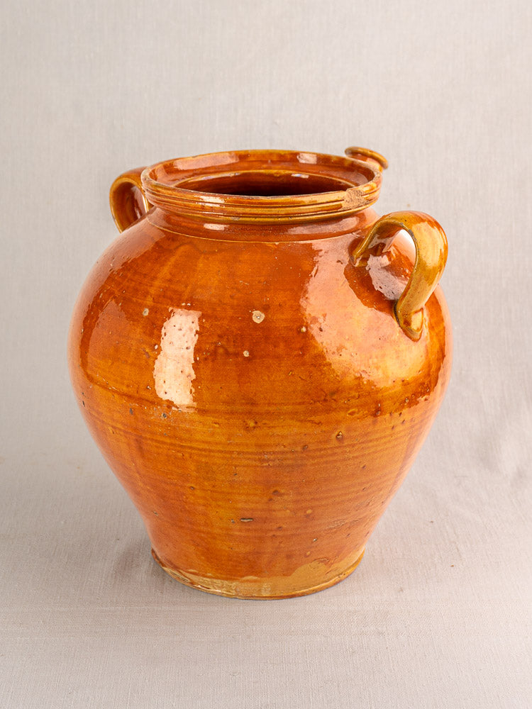 Antique French glazed earthenware cruche with honey-brown finish and double handles, on textured surface, neutral backdrop.