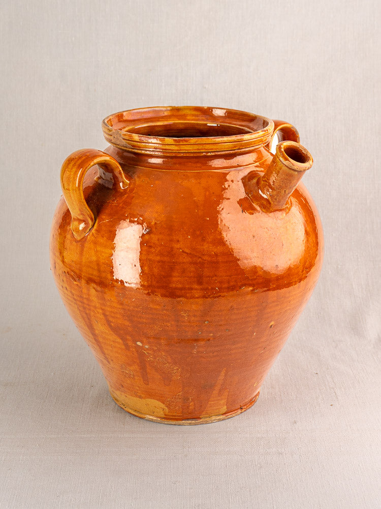 Antique French glazed earthenware cruche with honey-brown finish, two handles, and short spout on light gray surface.