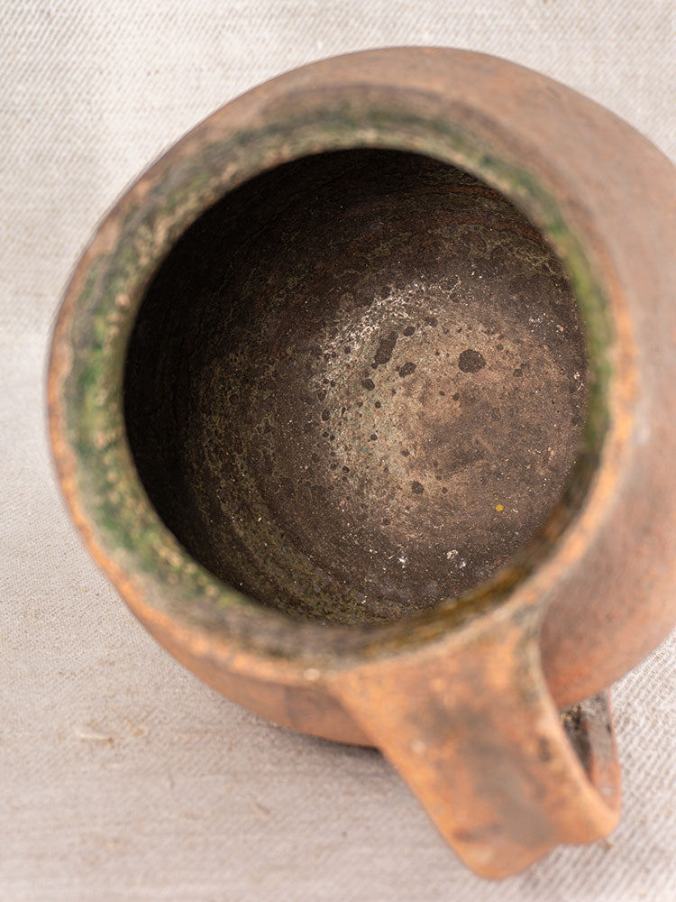 Antique French Toupin terracotta pitcher with green glaze and aged patina, handle detail on light textured fabric.