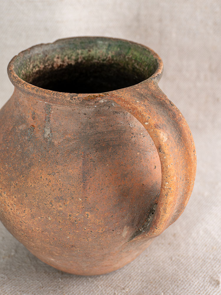Antique French terracotta pitcher with green glaze, textured brown and green surface on a neutral woven fabric background.