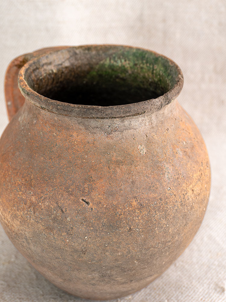 Antique French terracotta pitcher with green-glazed rim, wide opening, and single handle on light textured fabric.