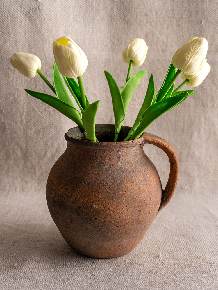 Antique French green-glazed terracotta toupin pitcher holding white tulips, on textured beige fabric background.