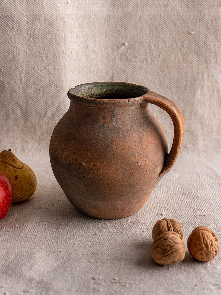 Antique French green-glazed terracotta toupin pitcher on beige fabric with apple, pear, walnuts; neutral background.