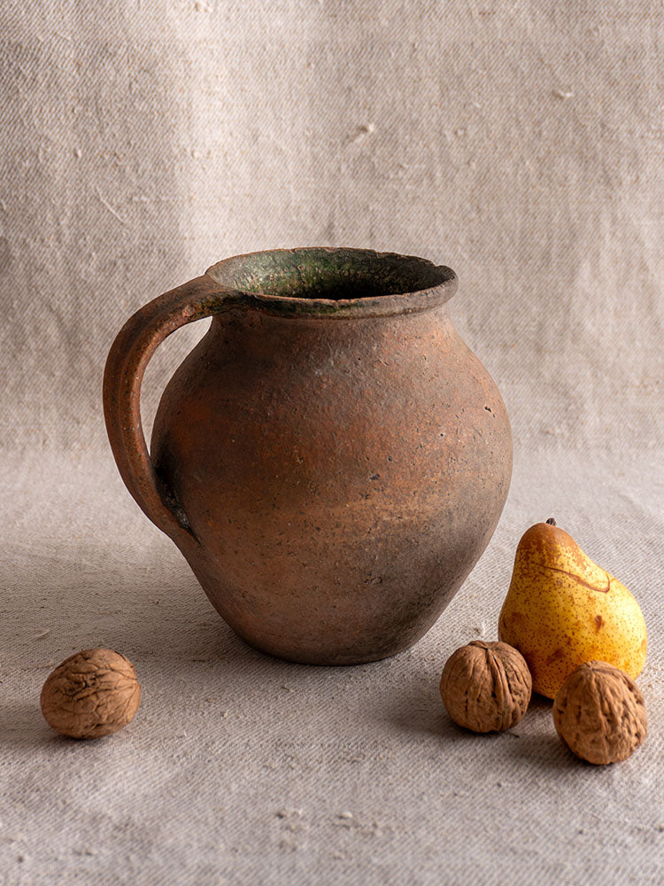 Antique French terracotta Toupin Provençal pitcher with green glaze beside a yellow pear and walnuts on textured fabric.