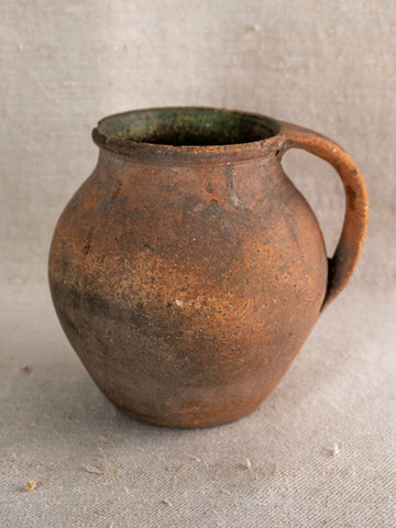 Antique French green-glazed terracotta pitcher with single handle on textured beige fabric, weathered from traditional use.