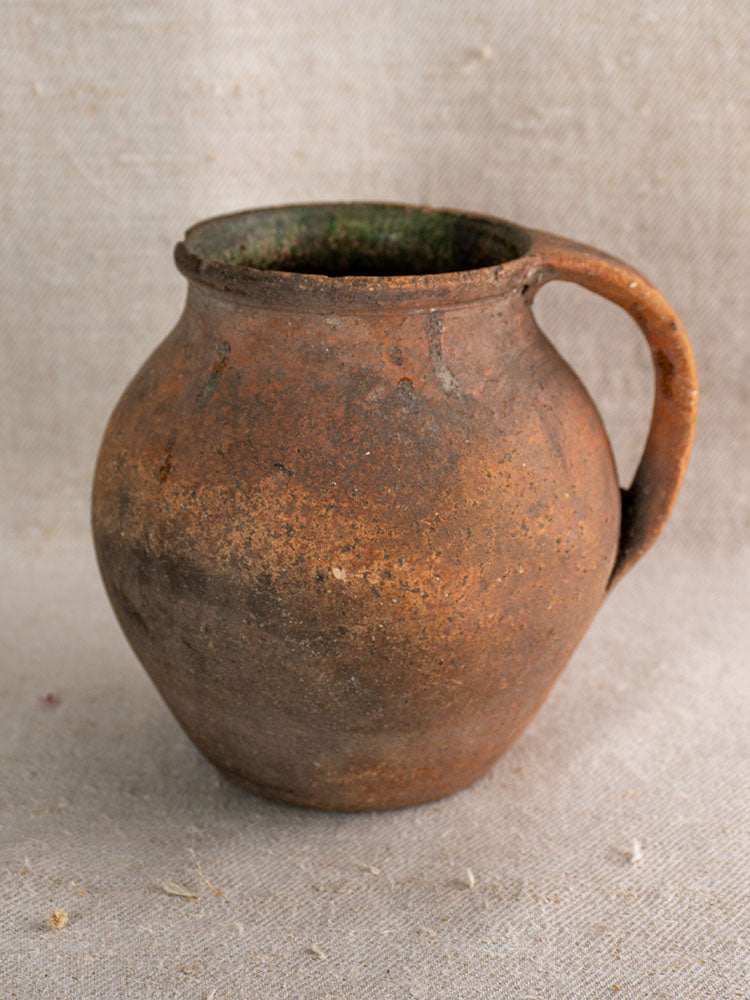 Antique French green-glazed terracotta pitcher with single handle on textured beige fabric, weathered from traditional use.