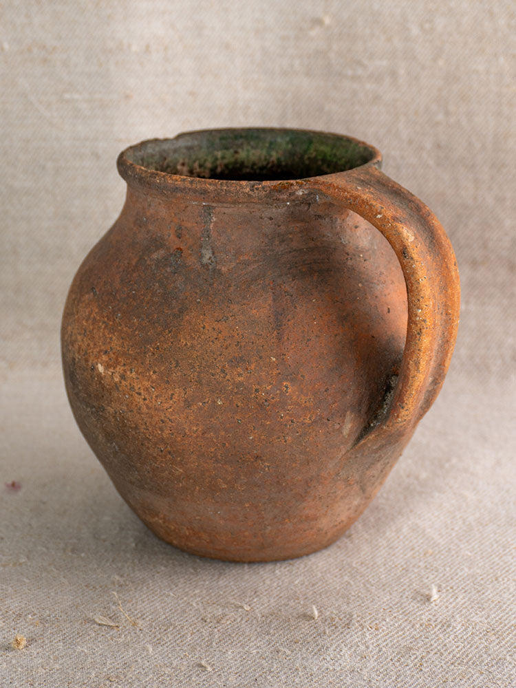 Antique French green-glazed terracotta toupin pitcher with single handle on textured beige fabric background.