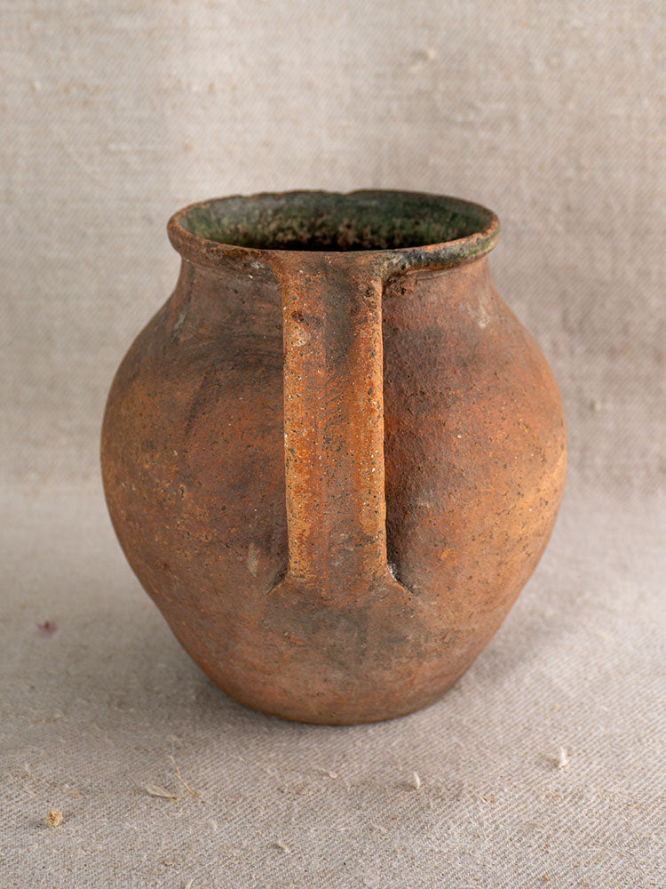 Antique French toupin terracotta pitcher with green glaze, patina, and thick handle on beige fabric.
