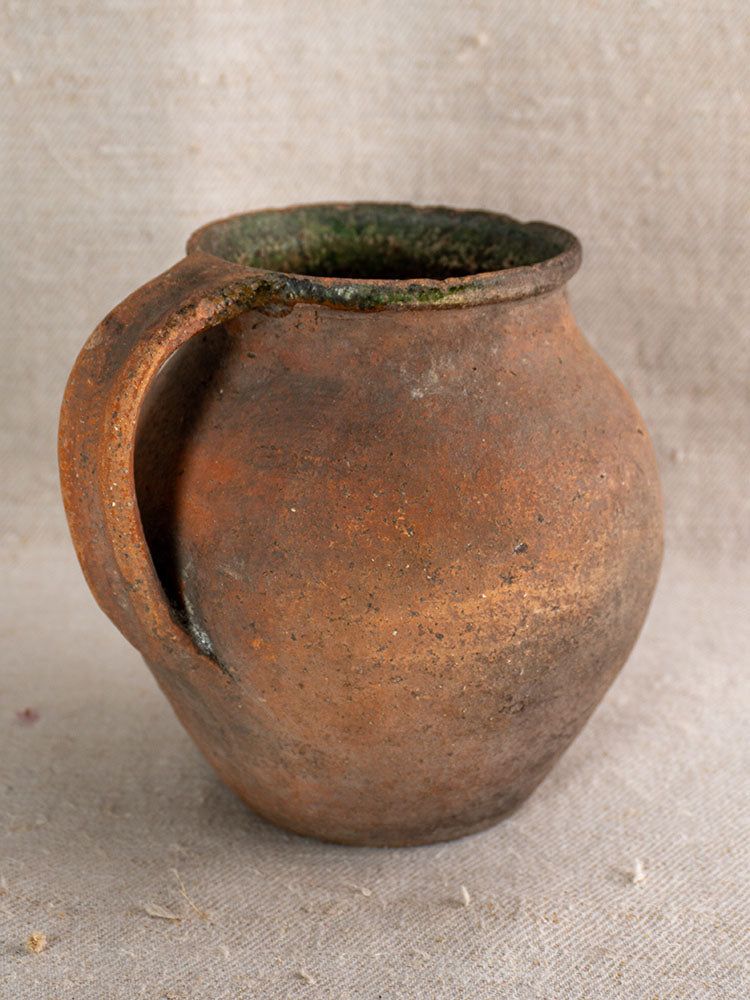 Antique French terracotta Toupin pitcher with green-glazed rim and handle on textured beige fabric.