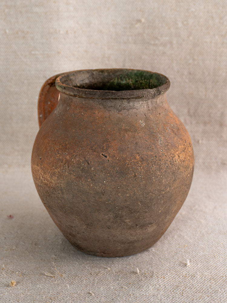 Antique French terracotta Toupin pitcher with green glaze and handle, aged patina, on light beige textured fabric.