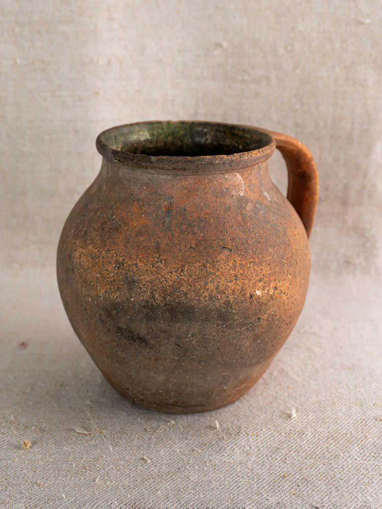 Antique French Toupin Provençal terracotta pitcher with green glaze and single handle on textured beige fabric, showing wear.