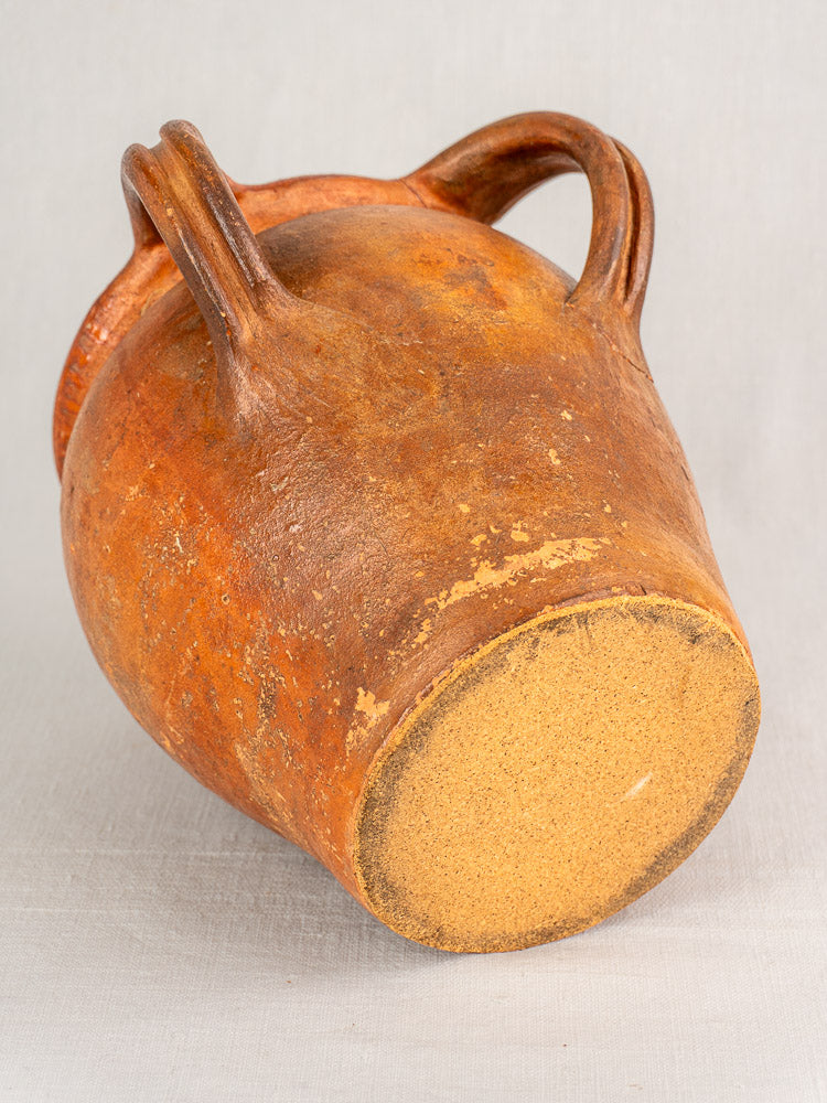 Antique French two-handle earthenware jug with orange-brown glaze and unglazed base on a pale background.