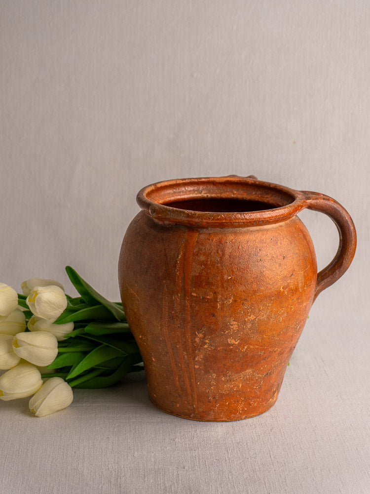 Antique French two-handle glazed earthenware jug with aged patina, displayed beside white tulips on neutral linen.