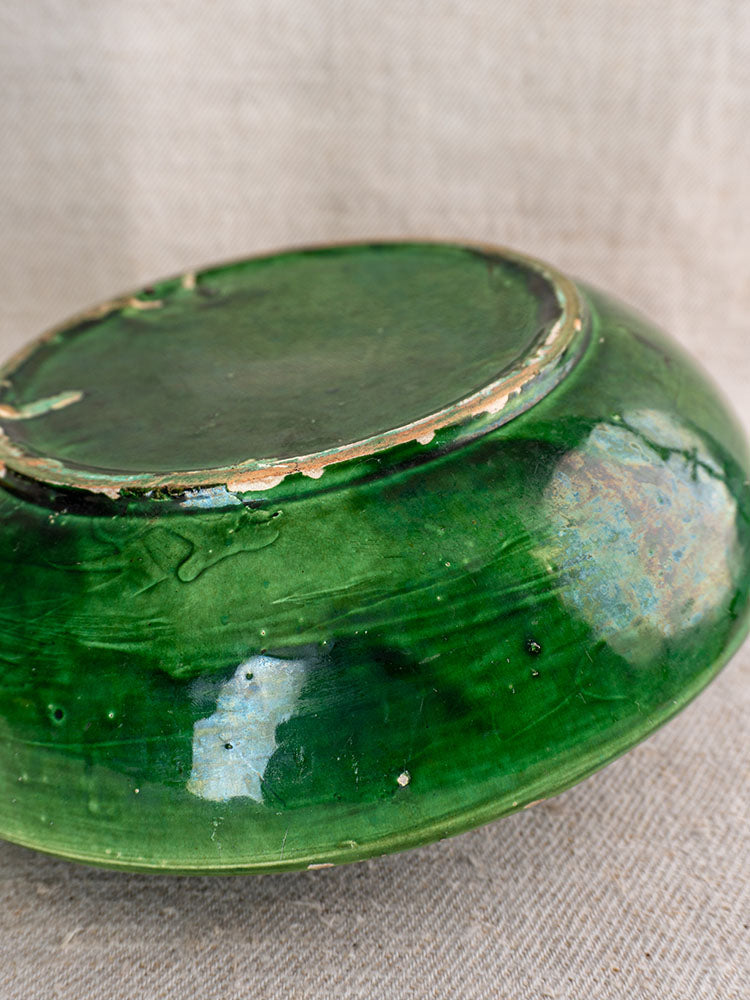 Underside of antique French green glazed earthenware bowl showing chips, wear, and exposed clay on beige fabric.