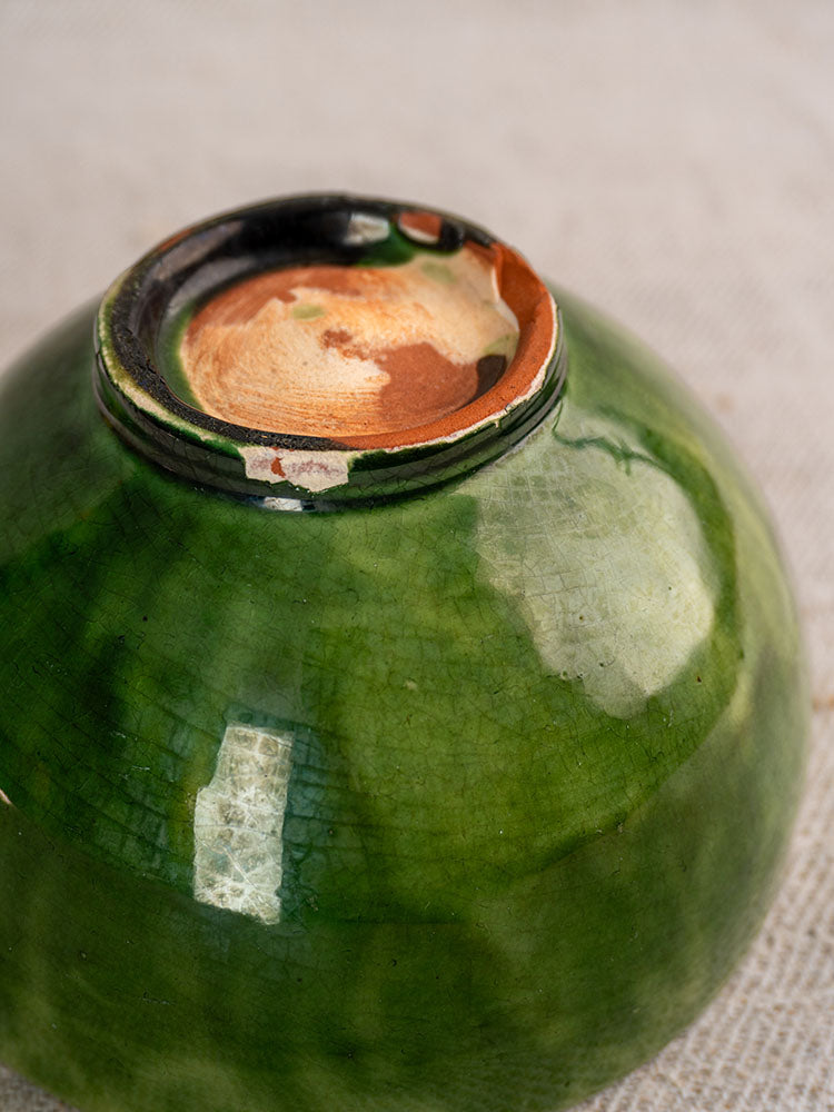 Underside of antique French green glazed earthenware bowl with chipped rim and exposed unglazed clay on light textured surface.