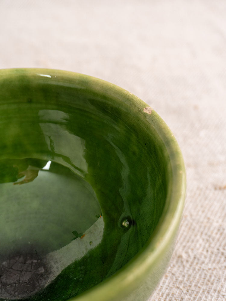 Antique French green glazed earthenware bowl with rim chip and partial water fill on textured beige fabric.