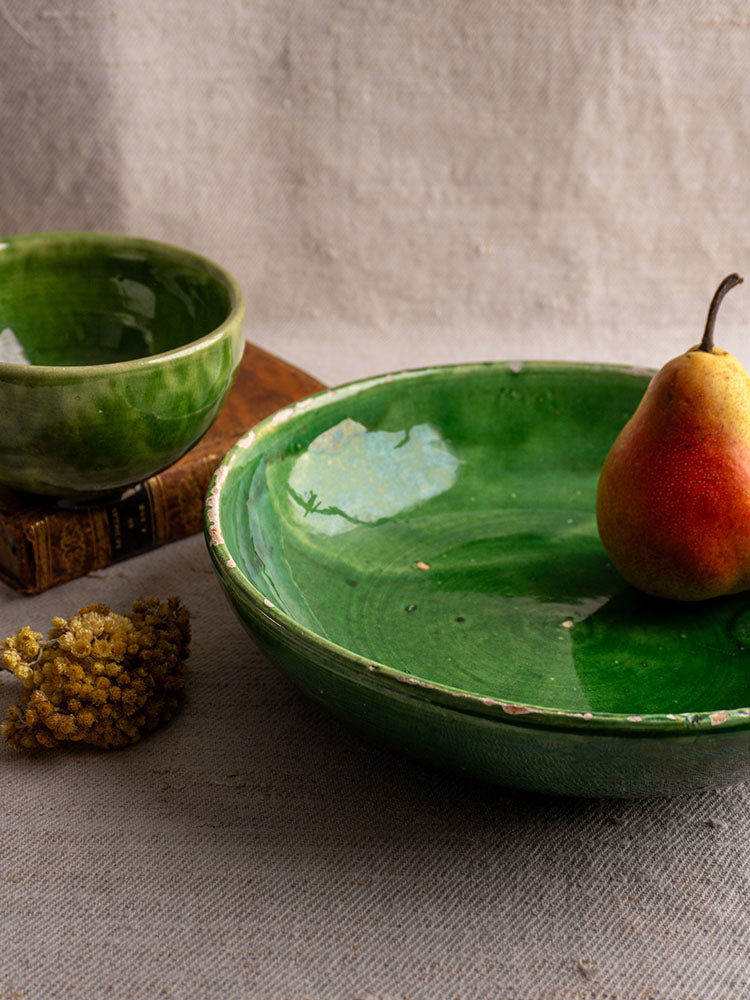 Antique French green glazed earthenware bowls set with pear, dried yellow flowers, and book on beige textured fabric.