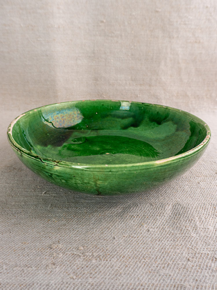 Antique French green glazed earthenware bowl with variegated glaze on textured beige fabric background.