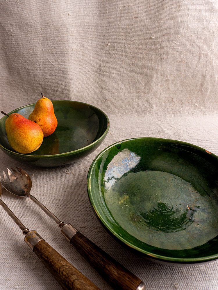 Green glazed vintage French ceramic serving bowls with pears, beside wooden utensils on beige textured fabric.