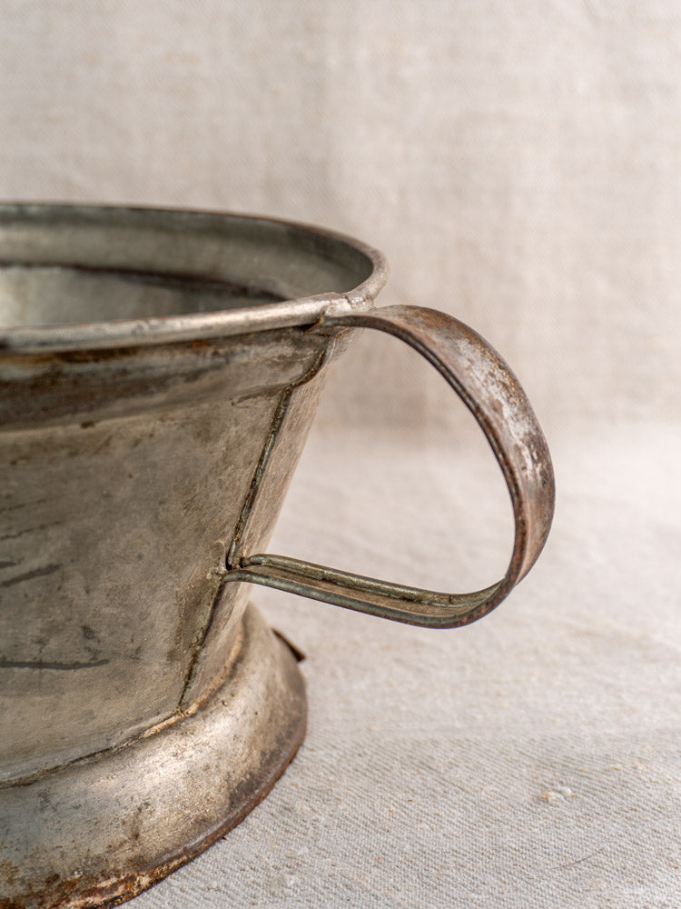 Antique French passoire in tinned sheet iron with pierced rosette base, handle shown against textured fabric.