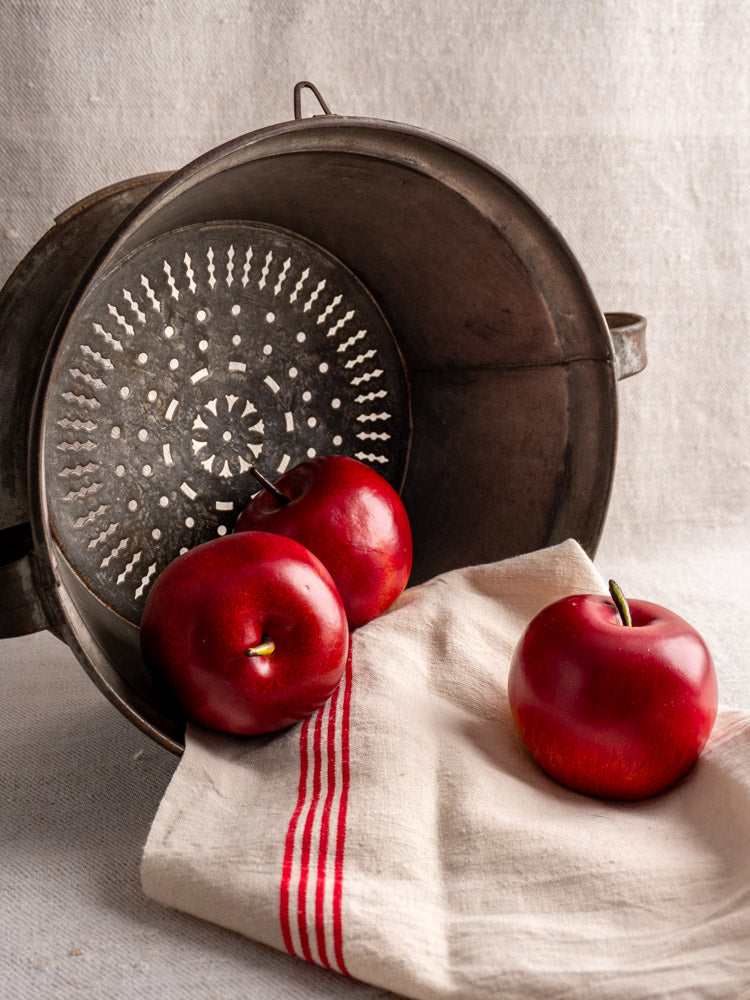 Antique French tinned sheet iron passoire with pierced rosette base beside three red apples on striped cloth.